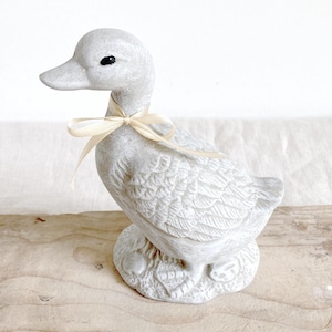 May include: A gray duck figurine with a cream-colored ribbon tied around its neck. The duck has detailed feather and wing patterns. The figurine is made of a stone-like material and sits on a base. The duck is a decorative item.