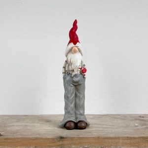 Può includere: Una statuetta di gnomo grigia e bianca con un cappello rosso e una canna di zucchero rossa e bianca in mano. Lo gnomo è in piedi su una superficie di legno.