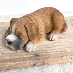 Könnte beinhalten: Eine braun-weiße Welpenfigur in ruhender Position. Der Welpe hat ein strukturiertes Fell, eine dunkle Schnauze und weiße Abzeichen an Gesicht und Pfoten. Er liegt mit dem Kopf auf den Vorderpfoten und zeigt eine detaillierte Fellstruktur.