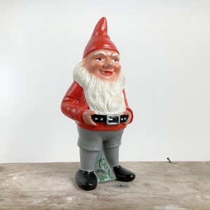 May include: A ceramic garden gnome figurine. The gnome wears a red pointed hat, a red jacket, gray pants, and black shoes. It has a white beard and a black belt. The figurine is standing on a wooden surface.