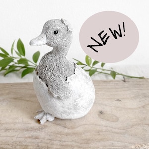 May include: A gray duckling figurine emerging from a cracked eggshell. The duckling has a textured body and a light gray beak. The word "NEW!" is in a pink circle. The figurine is on a wooden surface with green leaves.