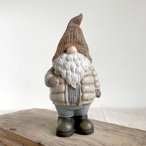 Può includere: Statuetta decorativa a forma di gnomo con berretto di lana marrone e giacca a vento beige. Lo gnomo ha una lunga barba bianca, un naso rosa e indossa pantaloni grigi e stivali verde scuro. La statuetta è su una superficie di legno.