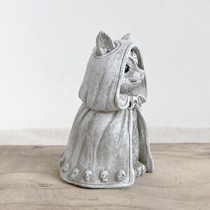 Puede incluir: Figura de gato gris con capa con capucha y detalles de calaveras. El gato est&aacute; sentado, con la cabeza girada hacia un lado. La figura tiene un aspecto envejecido, lo que sugiere una est&eacute;tica antigua o de &eacute;poca.