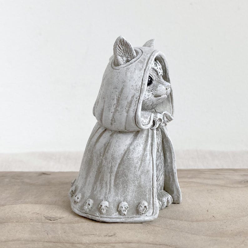 Puede incluir: Figura de gato gris con capa con capucha y detalles de calaveras. El gato est&aacute; sentado, con la cabeza girada hacia un lado. La figura tiene un aspecto envejecido, lo que sugiere una est&eacute;tica antigua o de &eacute;poca.