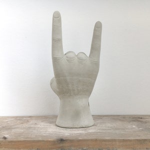 K&ouml;nnte beinhalten: Eine graue Betonskulptur einer Hand, die das 'Rock on'-Zeichen macht. Die Skulptur steht auf einer Holzoberfl&auml;che vor einer wei&szlig;en Wand. Die Hand zeigt detaillierte Kn&ouml;chel und eine strukturierte Oberfl&auml;che.