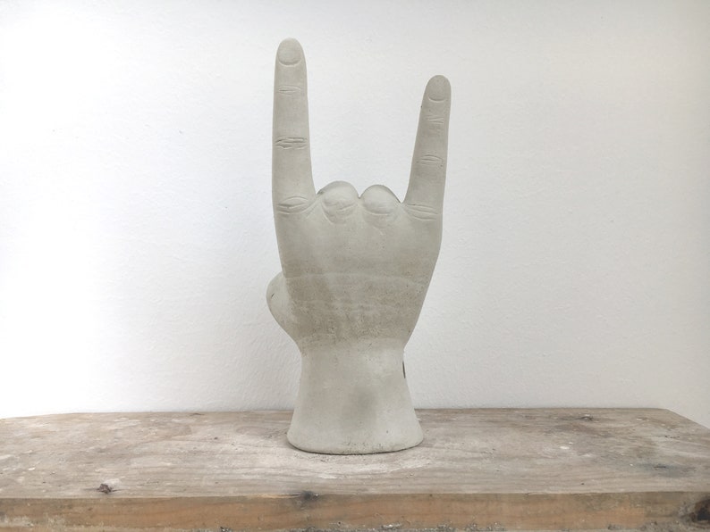 K&ouml;nnte beinhalten: Eine graue Betonskulptur einer Hand, die das 'Rock on'-Zeichen macht. Die Skulptur steht auf einer Holzoberfl&auml;che vor einer wei&szlig;en Wand. Die Hand zeigt detaillierte Kn&ouml;chel und eine strukturierte Oberfl&auml;che.