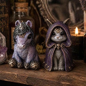 Puede incluir: Dos figuras de fantas&iacute;a: un unicornio morado con melena negra y un gato con una capa con capucha morada. Ambos tienen detalles dorados y pentagramas. Las figuras se exhiben en un estante de madera con una vela al fondo.