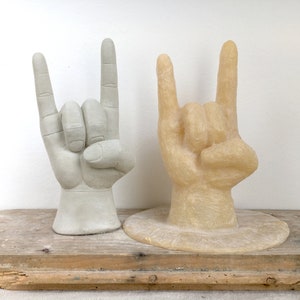 K&ouml;nnte beinhalten: Zwei Handskulpturen im 'Rock on'-Zeichen. Eine ist hellgrau, die andere hellbraun. Beide stehen auf einer verwitterten Holzoberfl&auml;che vor einem wei&szlig;en Hintergrund. Die Skulpturen sind wahrscheinlich Dekorationsartikel.