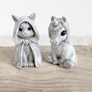 Puede incluir: Dos figuras grises: un gato con una capa con capucha y un unicornio. La figura del gato tiene una calavera en la capa y una estrella en la capucha. El unicornio tiene una estrella en la frente. Ambos tienen ojos oscuros y est&aacute;n sentados.