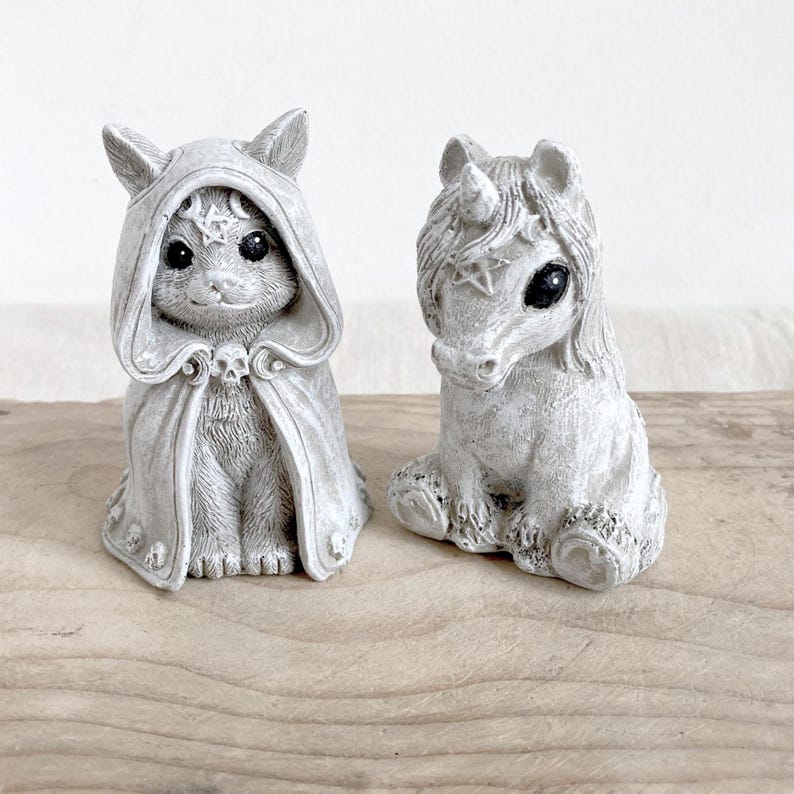 Puede incluir: Dos figuras grises: un gato con una capa con capucha y un unicornio. La figura del gato tiene una calavera en la capa y una estrella en la capucha. El unicornio tiene una estrella en la frente. Ambos tienen ojos oscuros y est&aacute;n sentados.