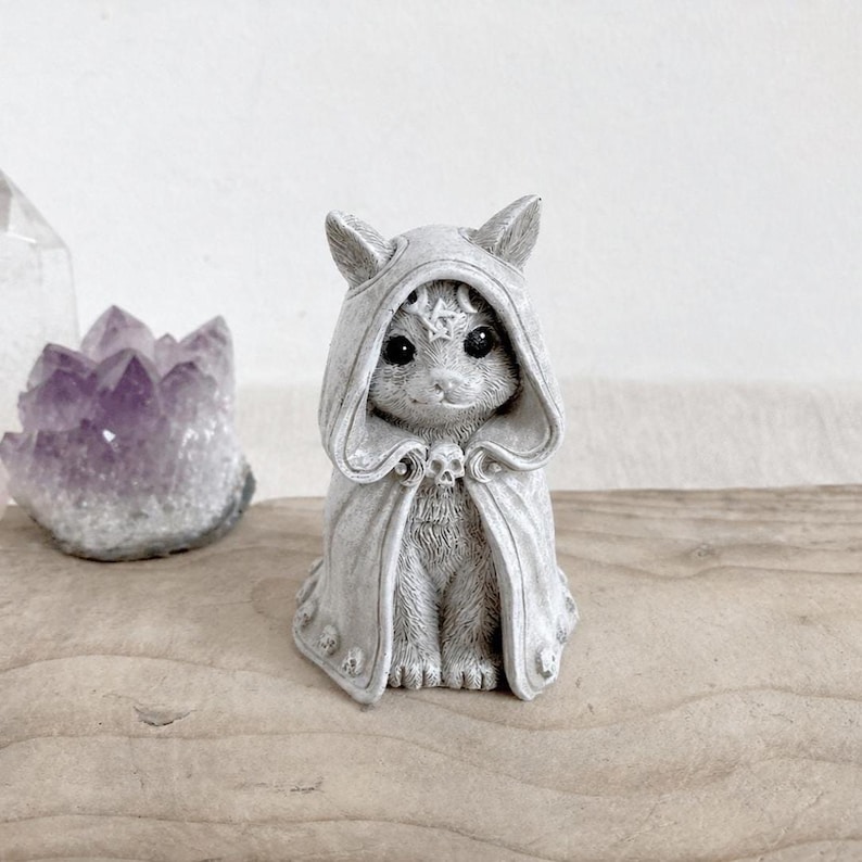 Puede incluir: Figura de gato gris con una capa con capucha con detalles de pentagrama y calavera. El gato est&aacute; sentado erguido, con ojos grandes y una textura de pelaje detallada. Un cristal p&uacute;rpura es visible en el fondo.