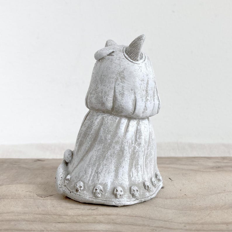 Puede incluir: Una figura gris con forma de gato con un acabado texturizado. El gato tiene orejas puntiagudas y una prenda drapeada. La base de la figura est&aacute; adornada con peque&ntilde;os detalles de calaveras, creando una est&eacute;tica g&oacute;tica &uacute;nica. La figura est&aacute; sobre una superficie de madera.