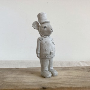 May include: A grey ceramic figurine of a mouse wearing a tall hat and a uniform. The mouse is standing on a wooden surface.