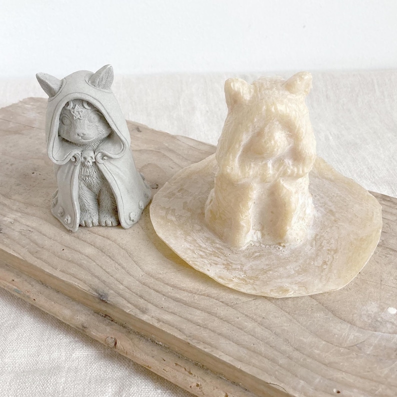 Puede incluir: Dos peque&ntilde;as figuras decorativas. Una es un conejito gris que lleva una capa con capucha con un emblema de calavera. La otra es una figura beige claro con forma de oso sobre una base circular. Ambas se exhiben sobre una tabla de madera.