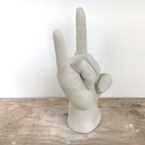K&ouml;nnte beinhalten: Eine hellgraue Skulptur einer Hand, die das 'Friedens-' oder 'Victory'-Zeichen macht. Die Finger sind nach oben ausgestreckt, wobei der Zeige- und Mittelfinger erhoben sind. Die Skulptur steht auf einer Holzoberfl&auml;che.