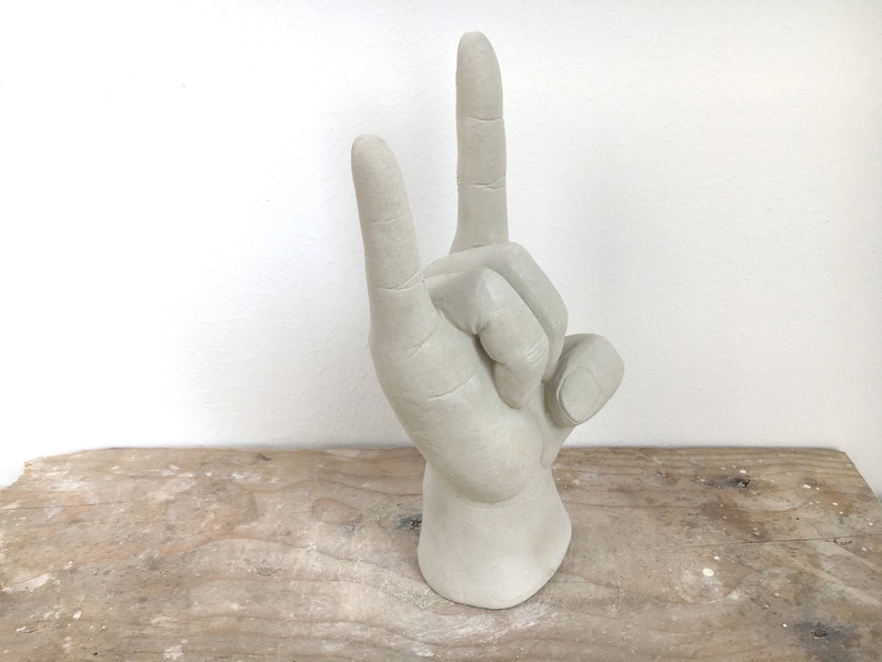 K&ouml;nnte beinhalten: Eine hellgraue Skulptur einer Hand, die das 'Friedens-' oder 'Victory'-Zeichen macht. Die Finger sind nach oben ausgestreckt, wobei der Zeige- und Mittelfinger erhoben sind. Die Skulptur steht auf einer Holzoberfl&auml;che.