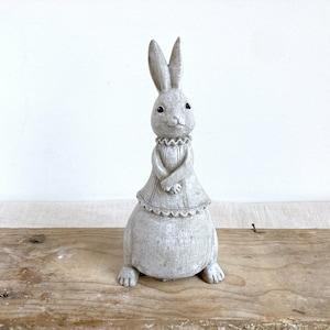 Könnte beinhalten: Eine graue Keramik-Hasenfigur mit einem weißen Kleid und einem Rüschenkragen. Der Hase steht auf einer Holzoberfläche.