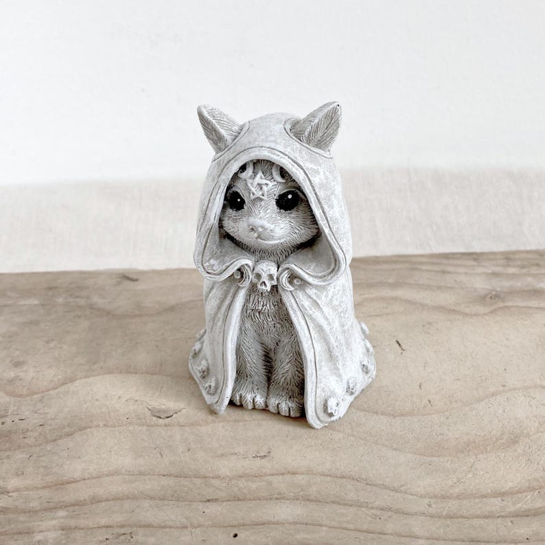 Puede incluir: Figura de gato gris con una capa con capucha y un broche de calavera. El gato tiene ojos grandes y un emblema de estrella en la capucha. La figura est&aacute; hecha de un material similar a la piedra y est&aacute; sobre madera.