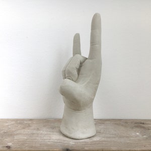 K&ouml;nnte beinhalten: Eine hellgraue Skulptur einer Hand, die die Geste &bdquo;Rock on&ldquo; ausf&uuml;hrt. Die Hand ist mit detaillierten Fingern und einer strukturierten Oberfl&auml;che geformt. Die Skulptur ist auf einem Sockel montiert und steht auf einer Holzoberfl&auml;che.