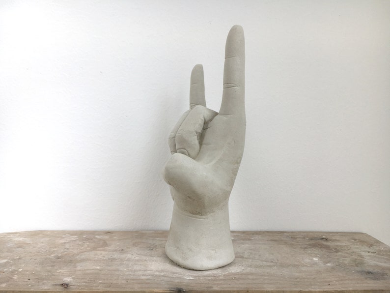 K&ouml;nnte beinhalten: Eine hellgraue Skulptur einer Hand, die die Geste &bdquo;Rock on&ldquo; ausf&uuml;hrt. Die Hand ist mit detaillierten Fingern und einer strukturierten Oberfl&auml;che geformt. Die Skulptur ist auf einem Sockel montiert und steht auf einer Holzoberfl&auml;che.