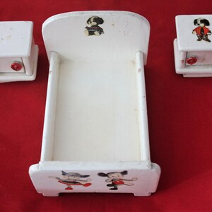 May include: A white wooden doll's bed with a white wooden headboard and footboard. The bed has a white mattress and is decorated with cartoon animal illustrations. The bed is accompanied by two white wooden bedside tables with red knobs.
