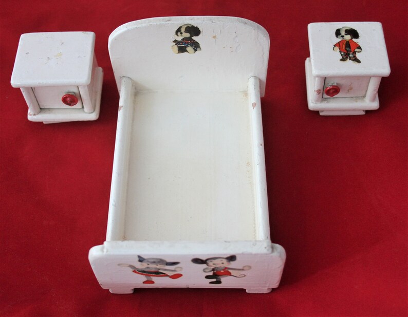 May include: A white wooden doll's bed with a white wooden headboard and footboard. The bed has a white mattress and is decorated with cartoon animal illustrations. The bed is accompanied by two white wooden bedside tables with red knobs.