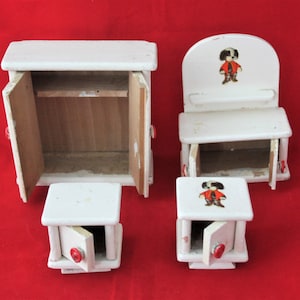May include: A set of four white wooden dollhouse furniture pieces. The set includes a cabinet with two doors, a chair with a back, and two small tables with drawers. Each piece has a red button on the front. The chair and one table have a printed image of a cartoon character wearing a red shirt and black trousers.