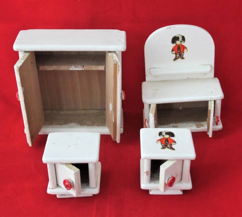 May include: A set of four white wooden dollhouse furniture pieces. The set includes a cabinet with two doors, a chair with a back, and two small tables with drawers. Each piece has a red button on the front. The chair and one table have a printed image of a cartoon character wearing a red shirt and black trousers.