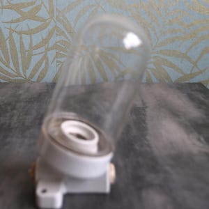 Könnte beinhalten: Eine klare Glaskuppel-Leuchte mit weißem Keramiksockel. Der zylindrische Glasschirm ist an einer runden weißen Halterung befestigt, die eine zentrale Fassung für eine Glühbirne aufweist. Die Leuchte enthält einen kleinen seitlichen Montagebügel.