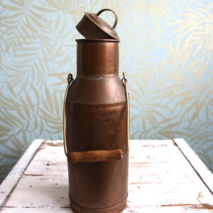 Könnte beinhalten: Eine hohe, zylindrische Milchkanne aus Kupfer mit einer verwitterten Patina. Sie verfügt über einen abnehmbaren Deckel mit einem kleinen, gebogenen Griff und einen stabilen Metallbügel mit einem glatten, horizontalen Holzgriff zum Tragen.