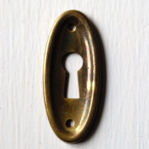 Op de afbeelding: Een ovale messing sleutelgatbedekking met een uitgesneden sleutelgat in het midden.