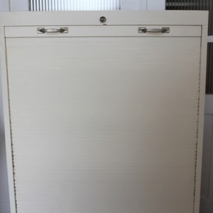 Könnte beinhalten: Weißer Metallschrank mit einer Rollladen-Tür und zwei silbernen Griffen. Der Schrank hat ein Schlüsselloch oben.