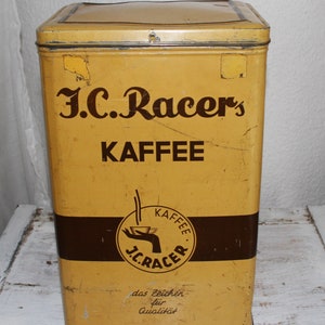 Könnte beinhalten: Eine gelbe Kaffeedose aus Metall mit dem Text "J.C. Racers Kaffee" und einem Logo einer Hand, die eine Tasse Kaffee hält, mit dem Text "J.C. Racers Kaffee" in einem Kreis. Der Text "das Zeichen fur Qualitat" ist unter dem Logo gedruckt.