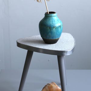 Op de afbeelding: Een kleine, grijze bijzettafel met een gespikkeld blad waarop een turquoise vaas met een gedroogde bloem staat. Een grote zeeschelp ligt eronder. De tafel heeft drie grijze poten en een metalen rand.