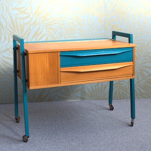 May include: A mid-century modern sewing table with a wooden top and drawers. The drawers are teal, and the legs are teal with small wheels. The cabinet is made of wood with a natural finish.