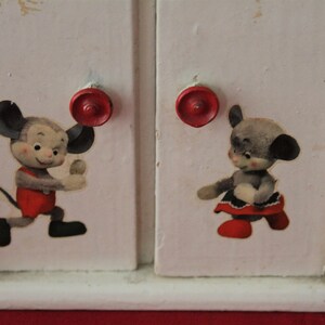 May include: White wooden toy cabinet with two doors and red knobs. Each door features a cartoon mouse illustration. One mouse is wearing a red shirt and the other is wearing a red dress.