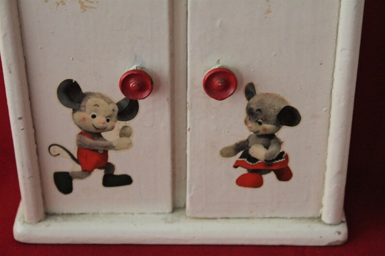 May include: White wooden toy cabinet with two doors and red knobs. Each door features a cartoon mouse illustration. One mouse is wearing a red shirt and the other is wearing a red dress.
