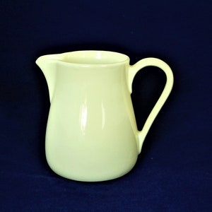 Op de afbeelding: Een lichtgele keramische kan met een rond lichaam en een handvat. De kan staat op een donkerblauwe achtergrond.