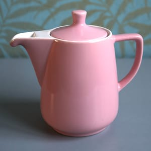 Könnte beinhalten: Eine rosa Keramik-Teekanne mit Deckel und Henkel. Die Teekanne hat einen Ausguss und einen weißen Akzent am Ausguss. Die Teekanne steht auf einer grauen Oberfläche.