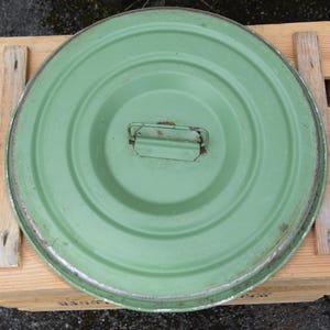 Op de afbeelding: Een ronde, lichtgroene metalen deksel met een handvat, met tekenen van slijtage. De deksel heeft een verhoogd midden en een lichtroestige rand. Het rust op een houten krat.