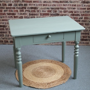 Könnte beinhalten: Ein hellgrün lackierter Holztisch mit einer Schublade und gedrechselten Beinen. Der Tisch ist etwa 76 cm lang und steht auf einem gewebten Jute-Teppich. Der Hintergrund ist eine Backsteinmauer.