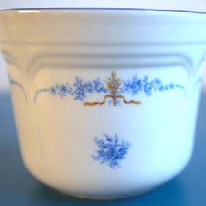 Könnte beinhalten: Eine weiße Keramiktasse mit blauem Rand und floralem Design. Das Design zeigt blaue Blumen und ein goldenes Band mit Weizenähren.