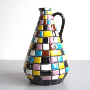 Op de afbeelding: Een kleurrijke keramische vaas met een geometrisch patroon van vierkanten in tinten blauw, geel, roze, bruin en zwart. De vaas heeft een handvat en een smalle hals.