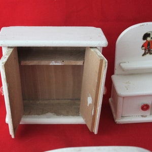 May include: Two white wooden dollhouse furniture pieces. The larger piece is a cabinet with two doors and a shelf inside. The smaller piece is a dresser with a shelf, two drawers, and a small dog illustration on the top.