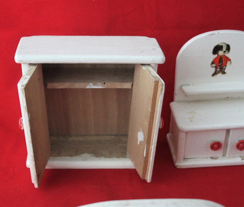 May include: Two white wooden dollhouse furniture pieces. The larger piece is a cabinet with two doors and a shelf inside. The smaller piece is a dresser with a shelf, two drawers, and a small dog illustration on the top.