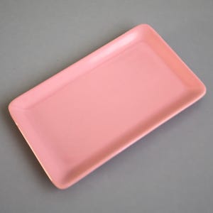 Könnte beinhalten: Ein rechteckiges, hellrosa Serviertablett mit abgerundeten Ecken. Das Tablett hat eine glatte, glänzende Oberfläche und ist vor einem grauen Hintergrund platziert. Dieses Tablett eignet sich zum Servieren von Speisen oder zum Präsentieren von Gegenständen.