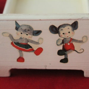 May include: A white wooden toy bed with two cartoon animals on the front. The animals are a grey cat with a red skirt and a grey mouse with red overalls. The bed has four legs.