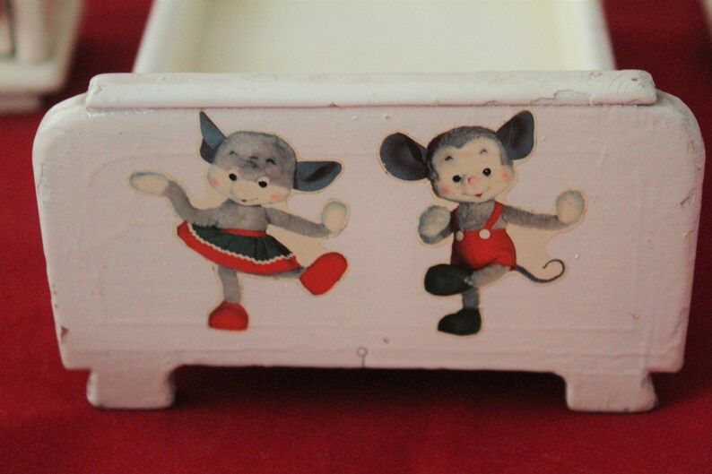 May include: A white wooden toy bed with two cartoon animals on the front. The animals are a grey cat with a red skirt and a grey mouse with red overalls. The bed has four legs.