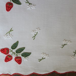 May include: White linen table runner with red embroidered strawberries and white embroidered flowers.