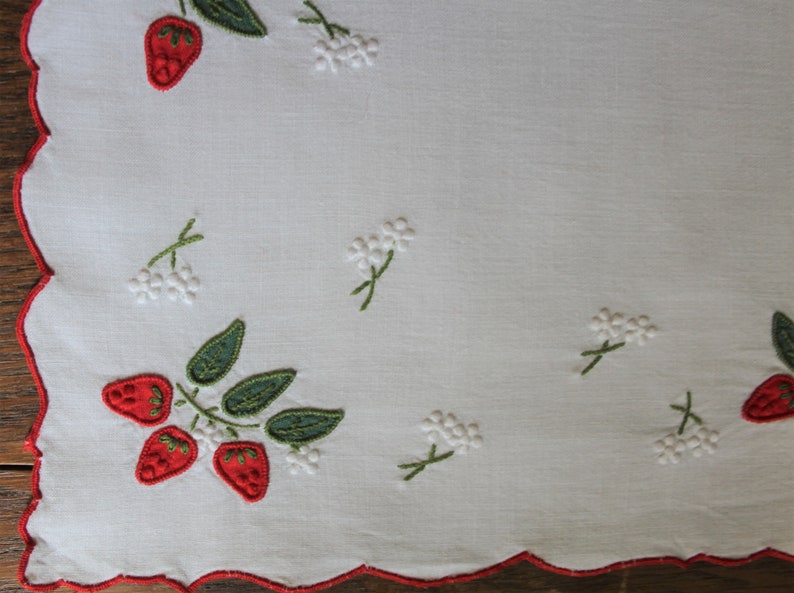 May include: White linen table runner with red embroidered strawberries and white embroidered flowers.